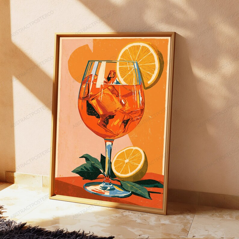 May include: A framed art print featuring a vibrant orange cocktail in a stemmed glass filled with ice cubes. A bright yellow lemon slice garnishes the glass, while another lemon slice and green leaves rest at the base on an orange surface.