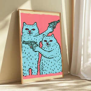 May include: A framed art print featuring two light blue cats with red eyes against a solid pink background. The cat on the left holds a revolver pointed upward, while the cat on the right holds a revolver pointed forward. The illustration uses bold black outlines.