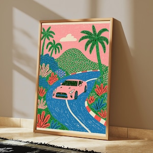 May include: A framed art print featuring a pink sports car driving along a winding blue road. The scene includes green palm trees, a textured green mountain, and a pink sky with a single white cloud. The road has red and white striped borders.
