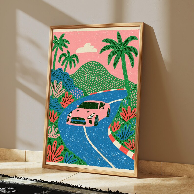 May include: A framed art print featuring a pink sports car driving along a winding blue road. The scene includes green palm trees, a textured green mountain, and a pink sky with a single white cloud. The road has red and white striped borders.