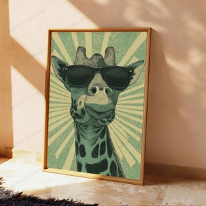 May include: A framed wall art print featuring a giraffe wearing black sunglasses. The giraffe is rendered in shades of green and cream with a sunburst background pattern. The artwork is enclosed in a simple wooden frame and displayed on a floor.
