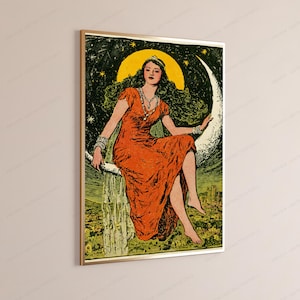 May include: A framed wall art print featuring a person with long dark hair wearing a vibrant orange dress and a silver necklace, sitting on a large white crescent moon against a dark green starry night sky above a landscape with buildings.