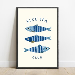 Pode incluir: Uma impress&atilde;o de arte emoldurada apresentando tr&ecirc;s peixes orientados horizontalmente com listras verticais azuis e azul-claras. O texto "BLUE SEA" est&aacute; impresso em azul escuro acima dos peixes e a palavra "CLUB" est&aacute; abaixo deles.