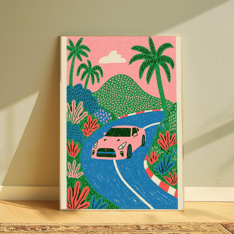 May include: A framed art print featuring a pink sports car driving along a winding blue road. The scene includes green hills, palm trees, and a pink sky with a single white cloud. The landscape is decorated with stylised red and green foliage.