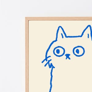 May include: A framed wall art print featuring a simple, hand-drawn illustration of a cat in vibrant blue ink on a cream-colored background. The cat design is rendered with thick, textured lines, showcasing large circular eyes and pointed ears.