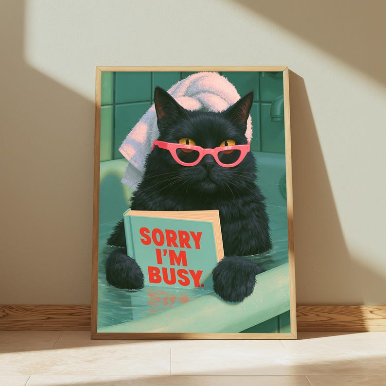 May include: A framed wall art print featuring a black cat wearing bright pink glasses and a white towel on its head while sitting in a green bathtub. The cat is holding a light blue book with the red text SORRY I'M BUSY printed on the cover.