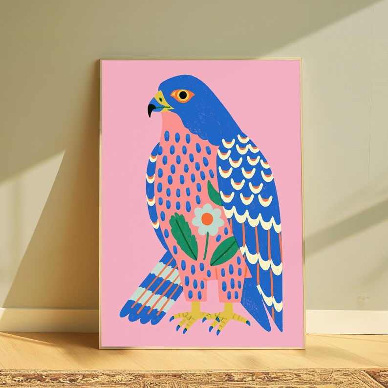 May include: A framed wall art print featuring a stylized blue falcon on a solid pink background. The bird has a yellow eye, beak, and feet, with a decorative white flower and green leaves on its chest and blue and white patterned wing feathers.