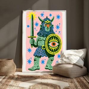 May include: A framed art print featuring a green, horned warrior figure on a pink background. The figure holds a grey sword and a yellow shield with a central eye motif. Blue star shapes are scattered across the pink field, and the warrior wears a textured blue tunic and helmet.