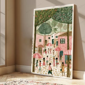 May include: A framed art print depicting a cobblestone street scene with pink buildings, green trees, and people walking under pink and white striped umbrellas. The illustration features a soft colour palette with various figures in casual clothing.