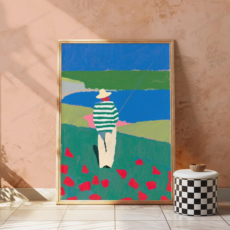 Op de afbeelding: Een ingelijste kunstdruk met een gestileerd schilderij van een persoon met een strooien hoed en een groen-wit gestreept shirt, staand in een veld met rode bloemen, uitkijkend over een blauw wateroppervlak onder een helderblauwe lucht.