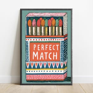 Op de afbeelding: Een ingelijste kunstdruk met een gestileerde illustratie van een open luciferdoosje gevuld met kleurrijke lucifers. Het centrale oranje rechthoekige paneel toont de witte tekst PERFECT MATCH. Het doosje is versierd met geometrische patronen in blauw, roze en geel.