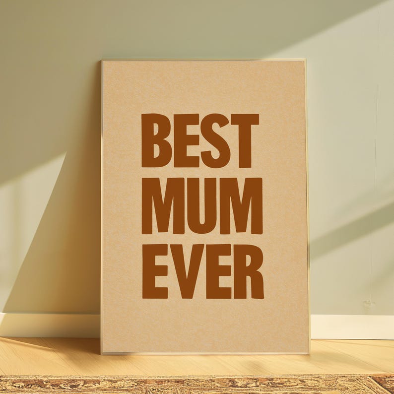 May include: A framed print with the words "BEST MUM EVER" in bold, brown letters. The print has a light brown background and is leaning against a wall. The frame is a light brown colour.