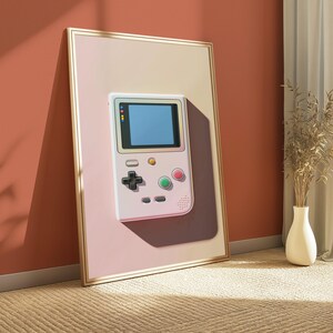 May include: A framed wall art print featuring a light-coloured handheld video game console with a blue screen, a black directional pad, and two circular buttons in green and pink, set against a soft gradient background within a thin gold frame.