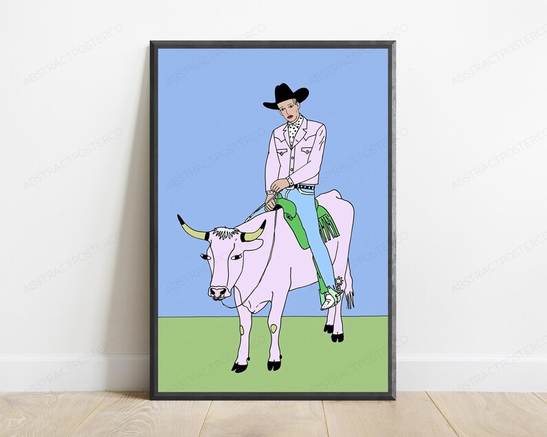 May include: A framed wall art print featuring a line-art illustration of a person wearing a black cowboy hat, a light pink jacket, and blue jeans, riding a light pink cow against a light blue sky and a solid light green ground.