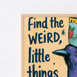 Op de afbeelding: Een ingelijste muurkunstprint met een lichtgele achtergrond en donkerblauwe handgeschreven tekst die Find the WEIRD, little things leest. Een deel van een paarse vogelillustratie met een kopje met bloemenpatroon op het hoofd is zichtbaar.