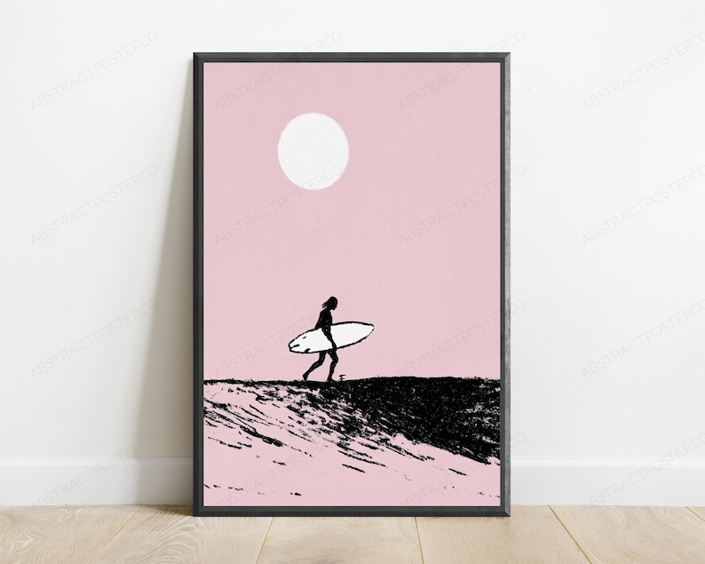 May include: A framed wall art print featuring a black silhouette of a person carrying a surfboard across a textured black landscape against a solid light pink background with a large white circle representing the sun positioned in the upper center.