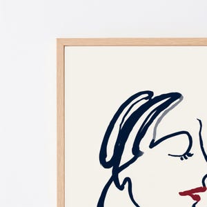 May include: A framed wall art print featuring a minimalist line drawing of a face in dark navy blue ink on a cream background, with a single bold red stroke highlighting the lips, enclosed in a light-toned wooden frame with a clean edge.