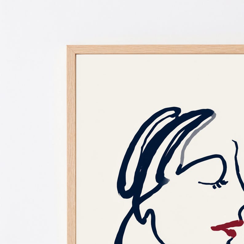 May include: A framed wall art print featuring a minimalist line drawing of a face in dark navy blue ink on a cream background, with a single bold red stroke highlighting the lips, enclosed in a light-toned wooden frame with a clean edge.