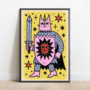 May include: A framed art print featuring a pink, horned character wearing chainmail armour and a black cape. The character holds a light blue sword and a pink shield with a red sun graphic. The background is yellow with black star symbols.