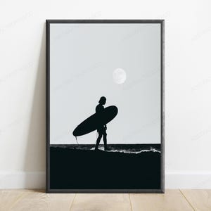 May include: A black and white framed wall art print featuring a silhouette of a person carrying a surfboard along the shoreline at night. A full moon is visible in the sky above the dark ocean waves and the sandy beach in this monochromatic scene.
