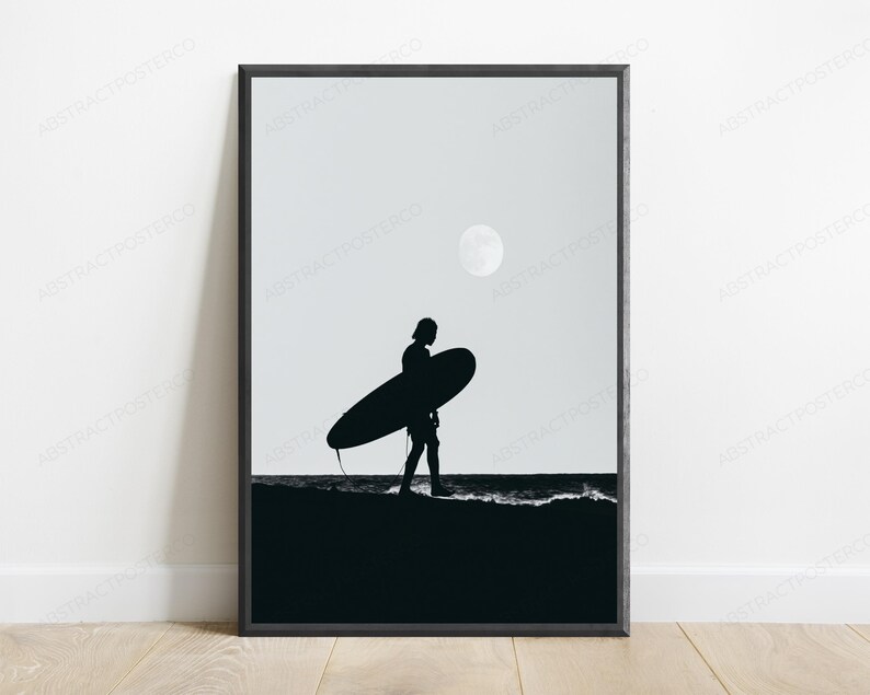 May include: A black and white framed wall art print featuring a silhouette of a person carrying a surfboard along the shoreline at night. A full moon is visible in the sky above the dark ocean waves and the sandy beach in this monochromatic scene.