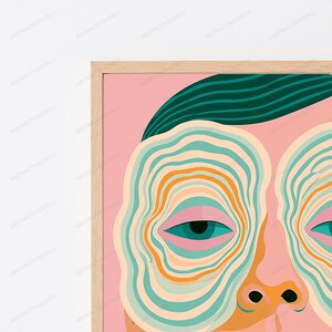 May include: A framed wall art print featuring a stylized face with a pink background. The face has dark teal hair, dark teal eyes, and concentric wavy lines in shades of teal, orange, and cream surrounding the eyes. The frame is light wood.