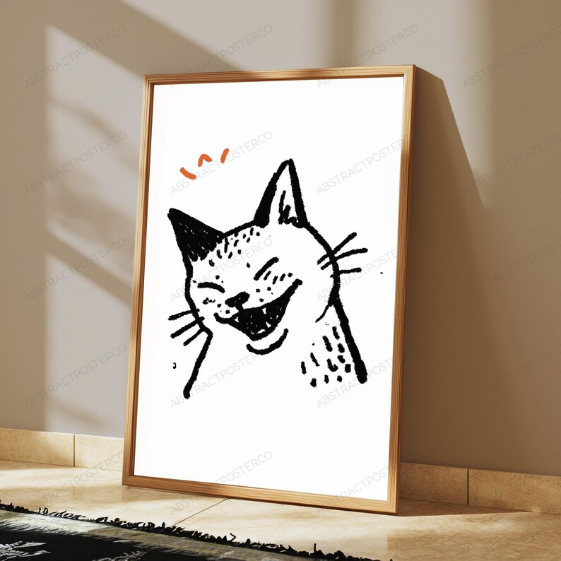 Pu&ograve; includere: Una stampa artistica incorniciata che presenta uno schizzo a inchiostro nero di un gatto che ride su sfondo bianco. Sopra la testa del gatto ci sono due piccoli segni curvi arancioni. L'opera &egrave; racchiusa in una sottile cornice dorata.