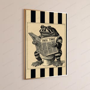 May include: A framed wall art print featuring a black and white illustration of a toad sitting on a toilet while reading a newspaper titled The Frog Times. The background consists of vertical black and beige stripes, and the print is encased in a thin gold frame.