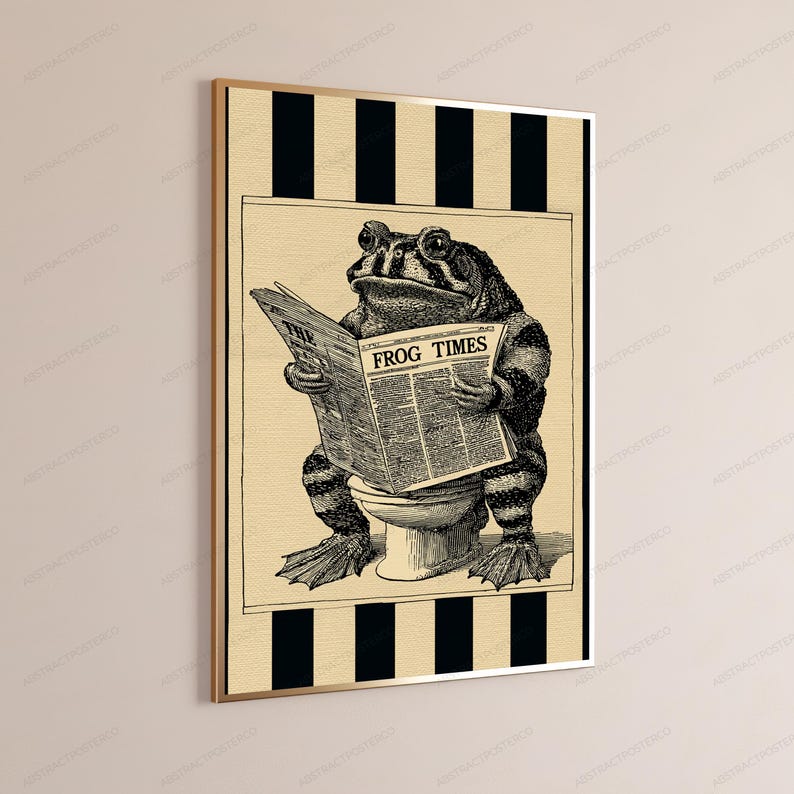 May include: A framed wall art print featuring a black and white illustration of a toad sitting on a toilet while reading a newspaper titled The Frog Times. The background consists of vertical black and beige stripes, and the print is encased in a thin gold frame.