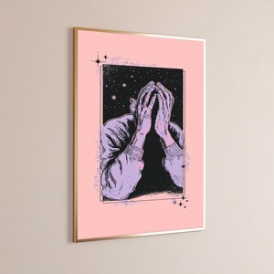 May include: A rectangular wall art print featuring a light pink background and a central black frame. Inside the frame, a stylized illustration shows a figure with light purple sleeves and hands covering their face against a starry night sky.