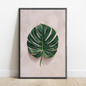 May include: A framed wall art print featuring a single, large, dark green monstera leaf centered against a pale, textured, light pink background. The frame is thin and black, providing a sharp contrast to the soft, muted tones of the botanical illustration.