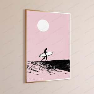 May include: A framed wall art print featuring a silhouette of a person carrying a surfboard across a dark, textured landscape against a solid light pink background with a large white circle representing the sun positioned in the upper center.