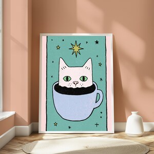 May include: A framed art print featuring a light pink cat with green eyes peeking out of a light blue coffee cup filled with black liquid. The background is a teal rectangle with small yellow stars and a large yellow star above the cat's head.