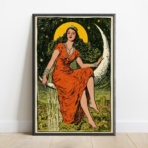 May include: A framed art print featuring a person with long dark hair wearing a vibrant orange dress and a tiara, sitting on a large white crescent moon against a dark green starry sky. Below the moon, a landscape with buildings is visible.