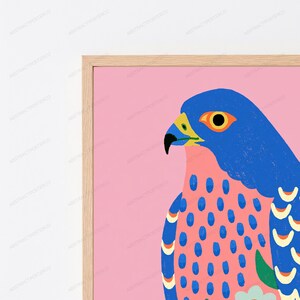 May include: A framed wall art print featuring a stylized blue falcon against a solid pink background. The bird has a bright yellow beak, a black eye, and a chest patterned with blue oval spots. The wings display blue and cream crescent shapes.
