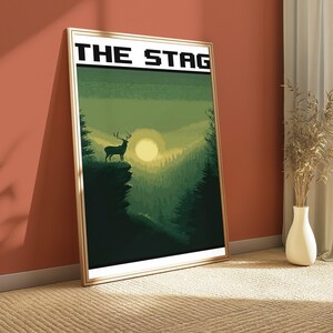 May include: A framed rectangular wall art print featuring a pixel art landscape with a stag standing on a cliff edge overlooking a forest at sunset. The text THE STAG is printed in a bold black pixelated font at the top of the white-bordered print.