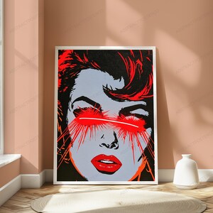 May include: A rectangular wall art print featuring a stylized graphic illustration of a face with grey skin, dark hair, and bright red lips. A horizontal white line crosses the eyes, with vibrant red light rays emanating outward from the eye area.
