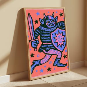 May include: A framed art print featuring a dark blue horned creature in armor holding a sword and a shield with a skull emblem. The background is a vibrant coral pink filled with scattered blue and black stars and small circles in a bold graphic style.