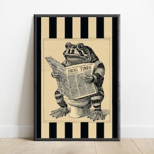 May include: A framed wall art print featuring a black and white illustration of a toad sitting on a toilet while reading a newspaper titled THE FROG TIMES. The central image is set against a background of vertical black and beige stripes.