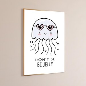 May include: A framed wall art print featuring a white cartoon jellyfish wearing pink heart-shaped sunglasses. The jellyfish has a smiling face with pink cheeks and wavy tentacles. Below the illustration, the text reads DON'T BE BE JELLY.