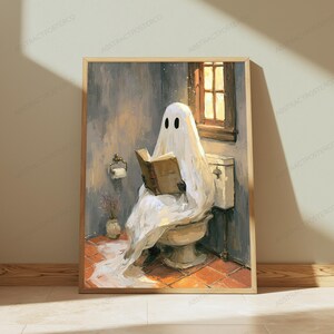 May include: A framed wall art print depicting a white ghost draped in a sheet sitting on a toilet in a bathroom, reading an open brown book. The scene features a window with warm light, a toilet paper holder, and a small vase on a tiled floor.