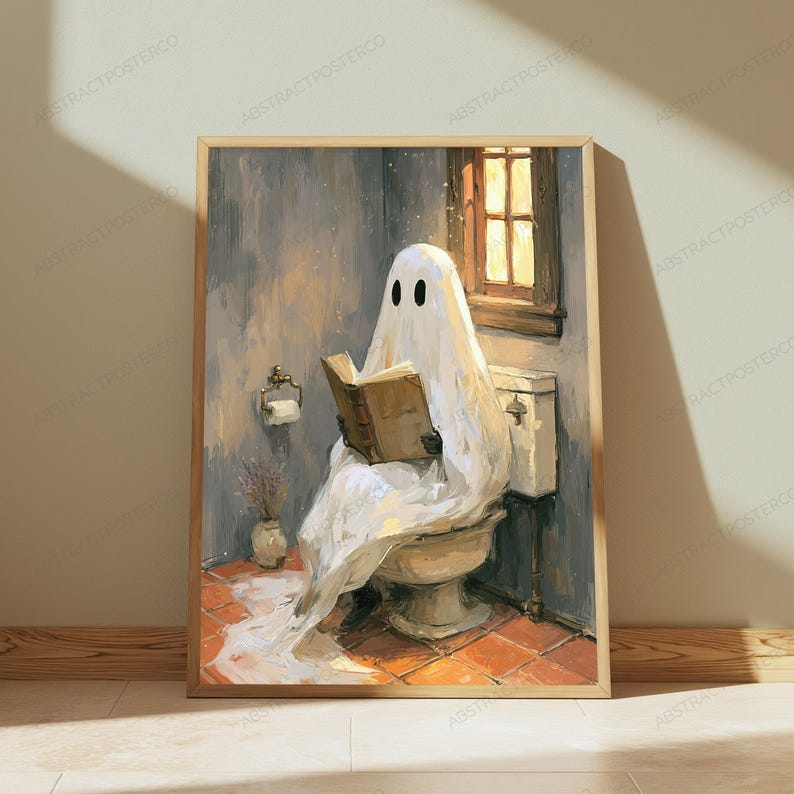 May include: A framed wall art print depicting a white ghost draped in a sheet sitting on a toilet in a bathroom, reading an open brown book. The scene features a window with warm light, a toilet paper holder, and a small vase on a tiled floor.