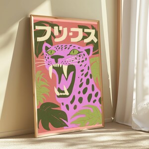 May include: A framed rectangular wall art print featuring a pink leopard with dark spots and an open mouth showing sharp teeth. The background is a salmon color with dark green tropical leaves and Japanese text at the top in cream-colored letters.