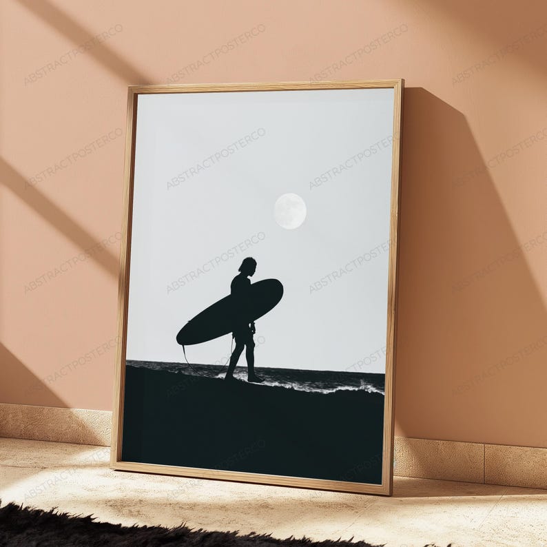 May include: A framed vertical wall art print featuring a black and white silhouette of a person carrying a surfboard on a beach. The scene includes a dark shoreline, a light sky, and a circular moon shape, all enclosed in a light wood frame.