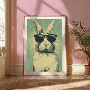 May include: A framed wall art print featuring a stylized illustration of a rabbit wearing black sunglasses. The rabbit is centered against a light green background with a radiating sunburst pattern. The artwork is enclosed in a thin gold frame.