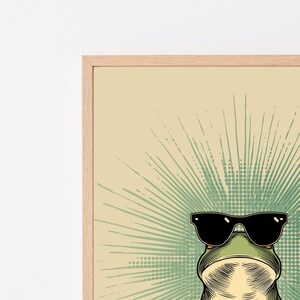 May include: A framed wall art print featuring a green frog wearing black sunglasses. The frog is centered against a light beige background with a radiating green sunburst pattern composed of lines and dots emanating from behind the frog's head.