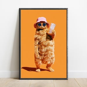 May include: A framed wall art print featuring an orange tabby cat standing upright against a solid orange background. The cat wears a light pink bucket hat and dark round sunglasses while holding a clear glass filled with ice cubes in one paw.