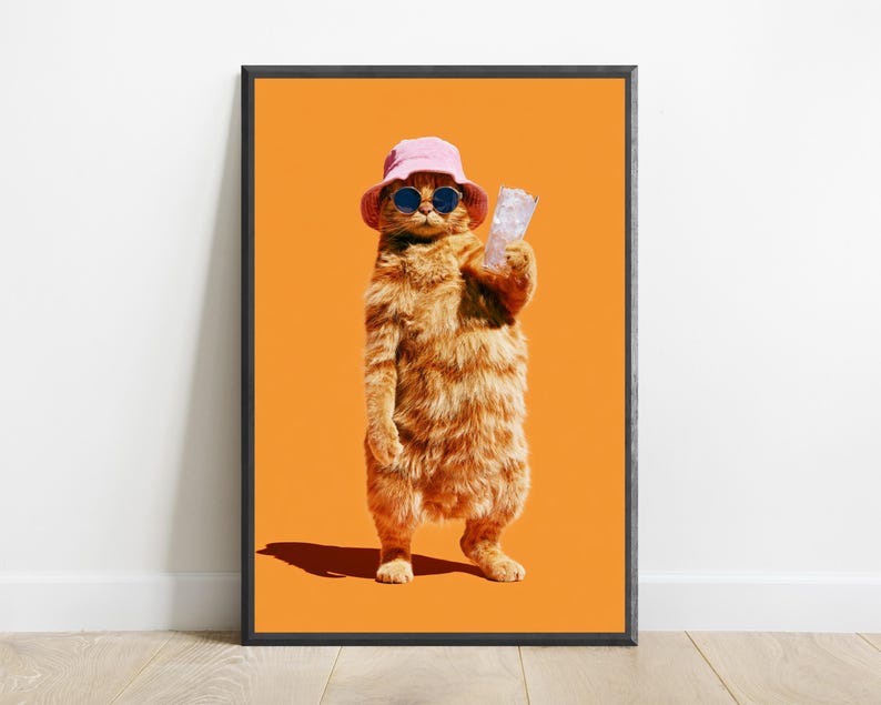 May include: A framed wall art print featuring an orange tabby cat standing upright against a solid orange background. The cat wears a light pink bucket hat and dark round sunglasses while holding a clear glass filled with ice cubes in one paw.