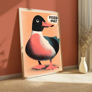 May include: A framed wall art print featuring a stylised illustration of a duck with a black head, pink chest, and dark wings against a peach background. A white speech bubble above the duck contains the black text "FEED ME!" in bold letters.