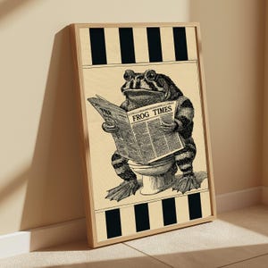 May include: A framed wall art print featuring a black and white illustration of a toad sitting on a toilet while reading a newspaper titled THE FROG TIMES. The background is a light cream colour with black vertical stripes at the top and bottom edges.